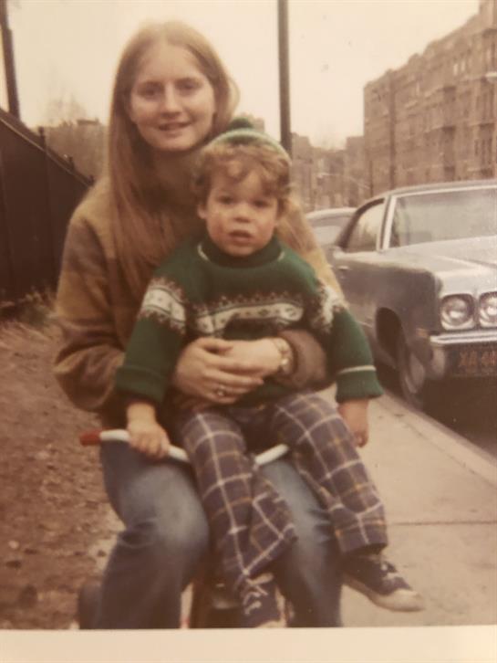 A woman sits on a sidewalk, holding a child, with a vintage car parked nearby in an urban area.