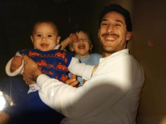 A father smiles while holding two children, creating a joyful moment in a cozy indoor environment.