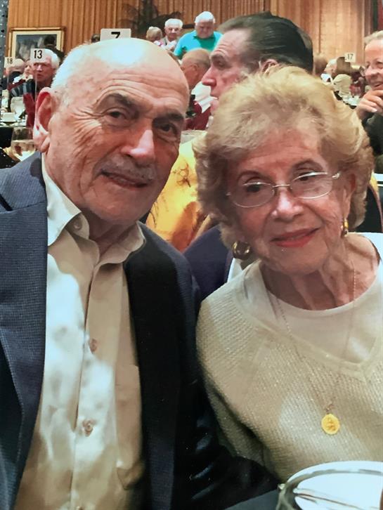 Two seniors sit closely together, smiling happily at a lively social event surrounded by friends.
