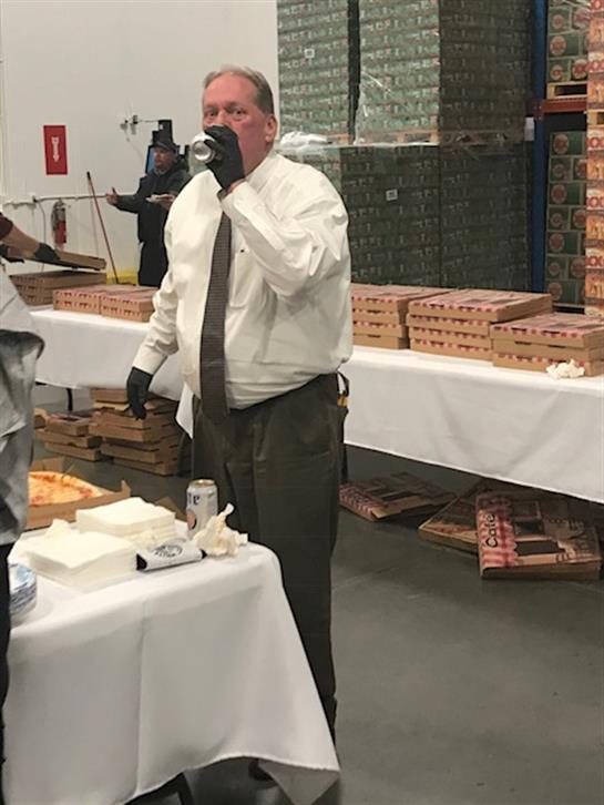 A businessman in formal attire holds a drink while interacting at a corporate event in a warehouse.