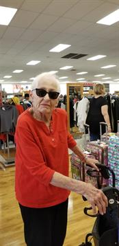 Senior person wearing sunglasses and a red shirt stands with a shopping cart in a crowded store.