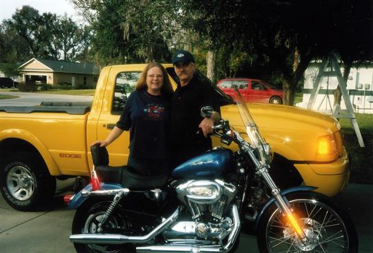 A smiling couple stands proudly next to a shiny motorcycle and a bright yellow truck in daylight.