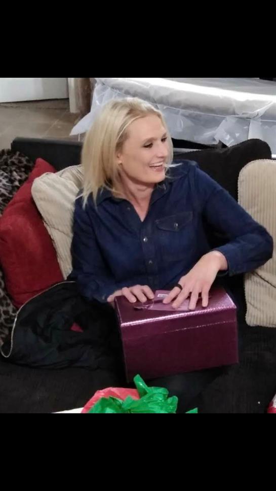 Woman smiles as she unwraps a gift while sitting on a comfortable couch indoors.