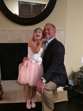 A girl wearing a pink dress is laughing and posing with a smiling adult near a fireplace.