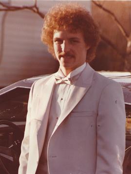 Man in a white suit stands confidently next to a vintage car during an outdoor event in the 1980s.