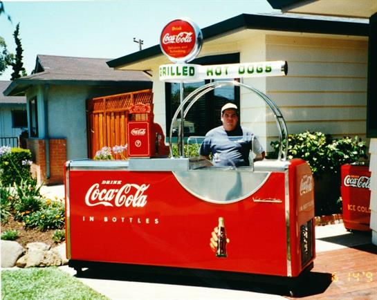 A vendor stands at a retro hot dog cart with Coca-Cola signage, serving customers outside a house.