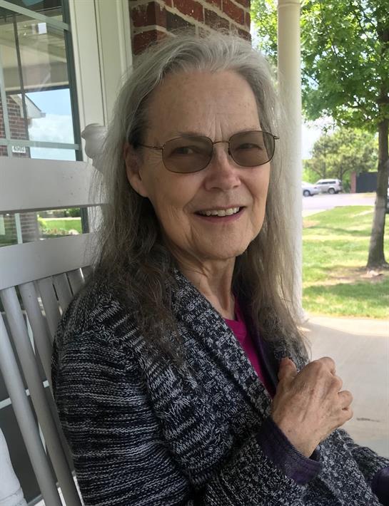 A woman with long gray hair and glasses sits on a porch, smiling and looking relaxed in the sun.