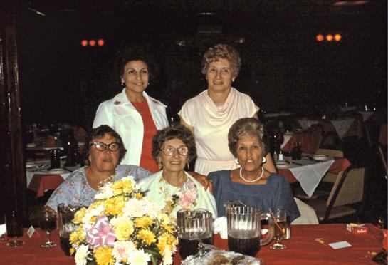 Five elderly women pose together at a festive gathering with flower arrangements and drinks.