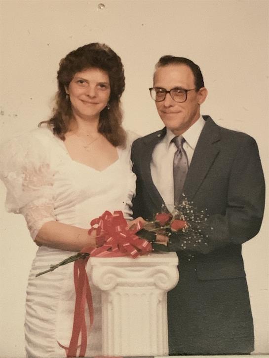 A couple in formal wear stands proudly together by flowers and a decorative pedestal.