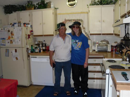 Two friends stand together in a cheerful kitchen, both wearing hats and smiling happily.