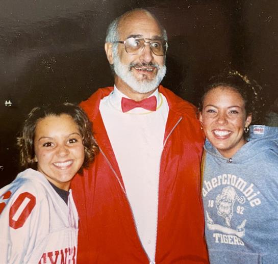 Two friends smile beside an entertainer in glasses and a red outfit at an evening event.