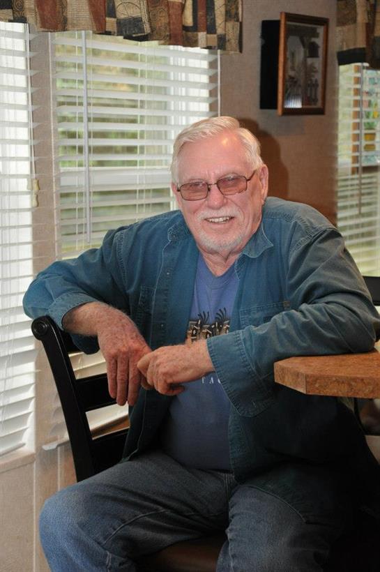 A smiling elderly man sits at a wooden table, relaxing in a sunlit cafe, enjoying his day.