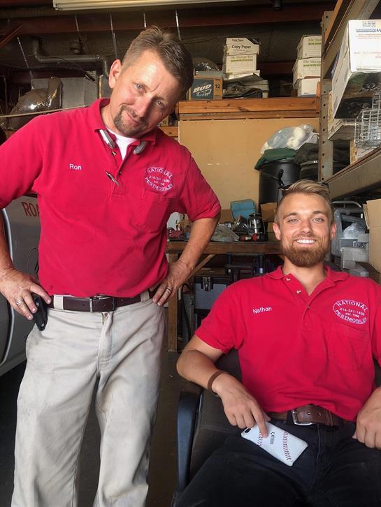 Two men smile and pose inside a workshop filled with tools and equipment while wearing red shirts.