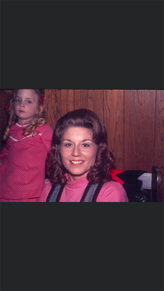 A joyful woman with curled hair sits next to a girl in a pink dress, both smiling in a cozy room.