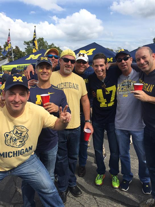 Friends gather at a tailgate party, wearing Michigan gear and enjoying refreshments in the sun.
