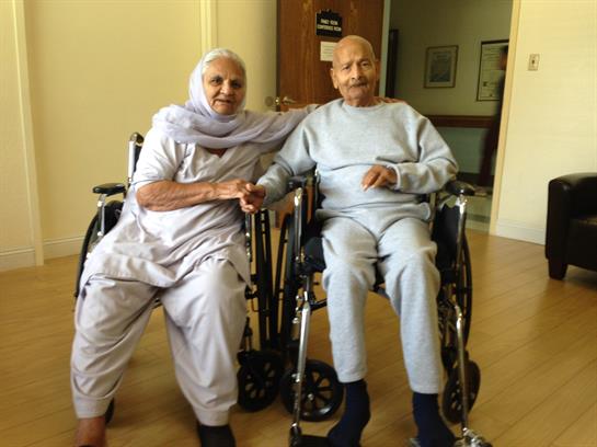 A loving elderly couple sits together in wheelchairs, smiling warmly and holding hands.