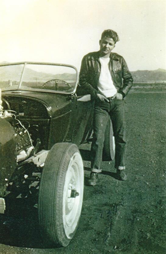 Young man in leather jacket leans against classic car under warm sunlight in open field.