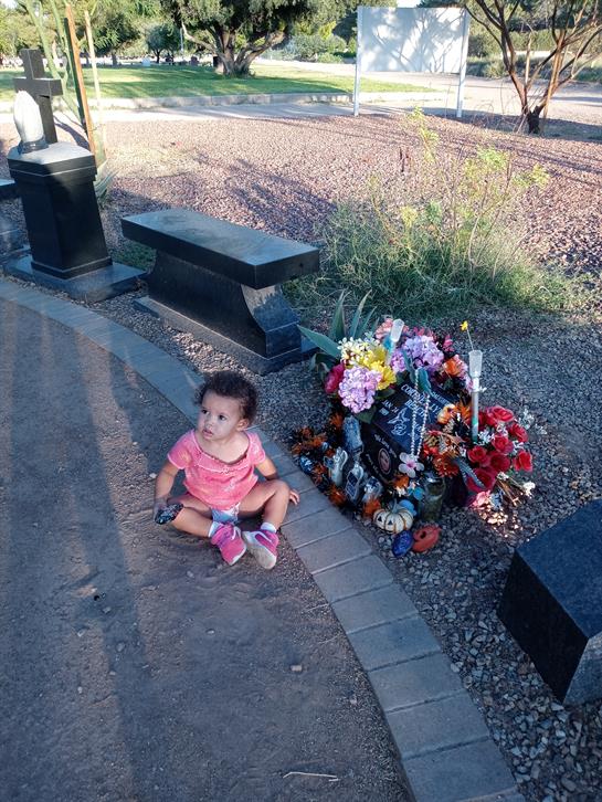 A young child sits on the ground beside a flower-decorated grave, reflecting on memory.