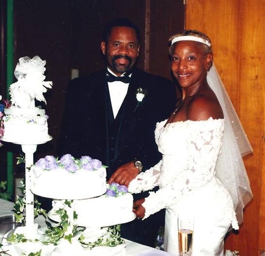 Newlyweds smile beside a floral cake at their wedding reception.