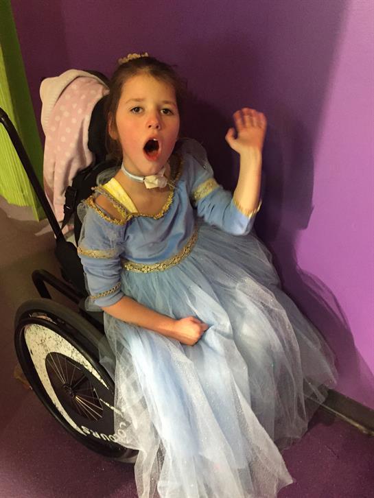 Girl in a blue dress waves with excitement while sitting in a wheelchair at a lively event.