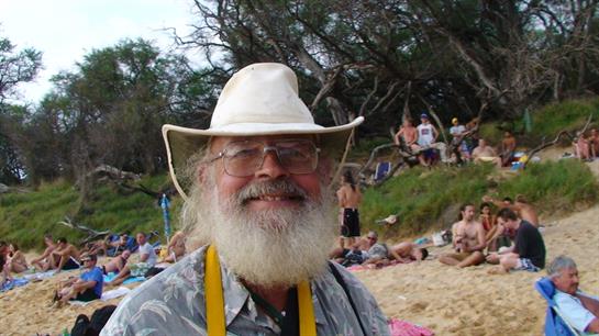 A cheerful man wearing a white hat and glasses smiles at the beach surrounded by people.