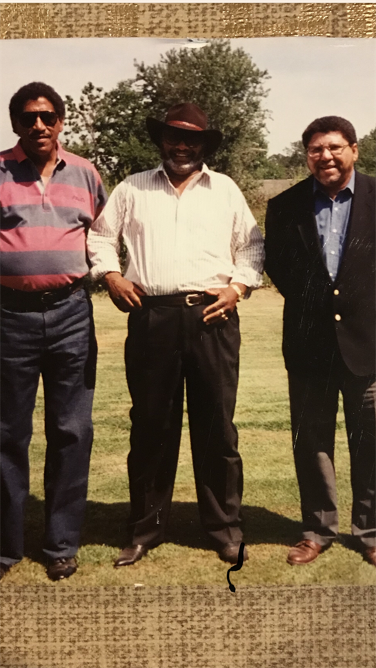 Three men pose together in a green park, smiling and enjoying a sunny afternoon outdoors.