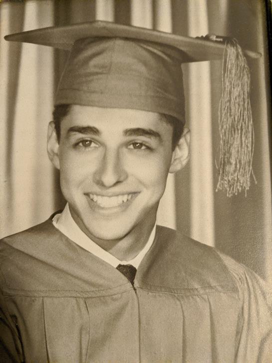 A young man in a graduation cap and gown smiles proudly while posing indoors during the celebration.