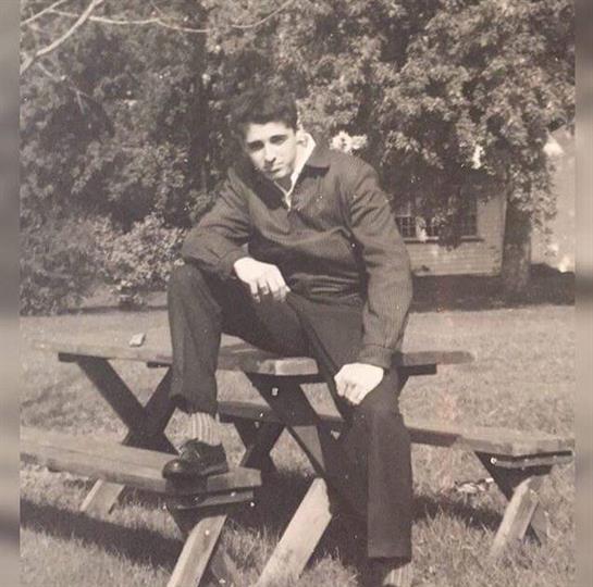 Young man sits relaxed on a picnic table, enjoying a sunny day in a rural park with greenery.