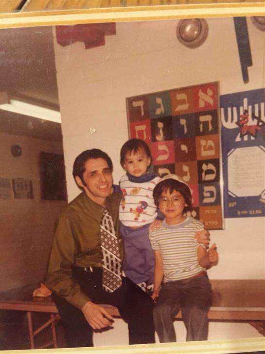 A father smiles with his two children in a colorful classroom filled with learning materials.