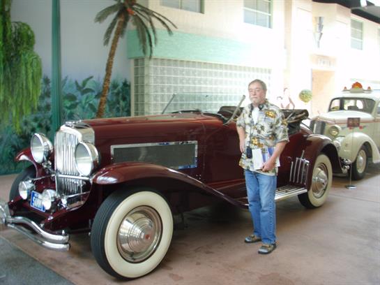 A man stands next to a vintage convertible car in a sunny coastal venue adorned with tropical decor.