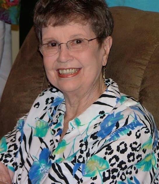 A cheerful elderly woman sits comfortably in her home, wearing a vibrant blouse.