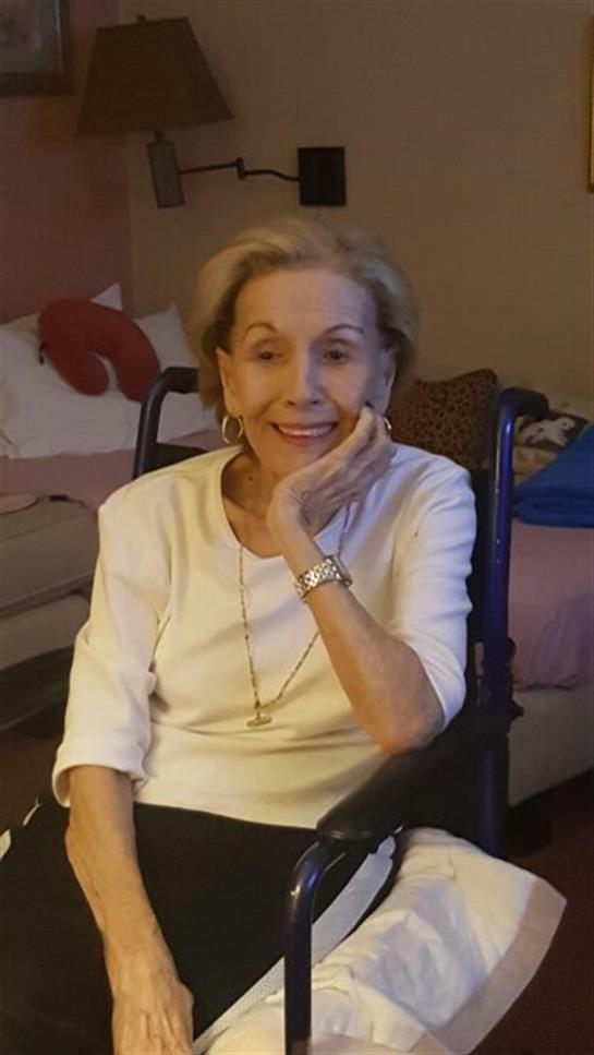 An elderly woman sits in a wheelchair, smiling and resting her chin on her hand in her cozy home.
