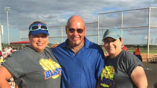 Three individuals smile together, showcasing camaraderie during a recreational softball event.