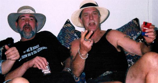 Two men sit together on a couch wearing casual clothing and hats, sharing drinks and laughter.