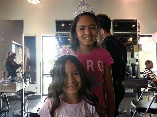 Two young girls stand together in a salon, smiling and wearing crowns for a party.