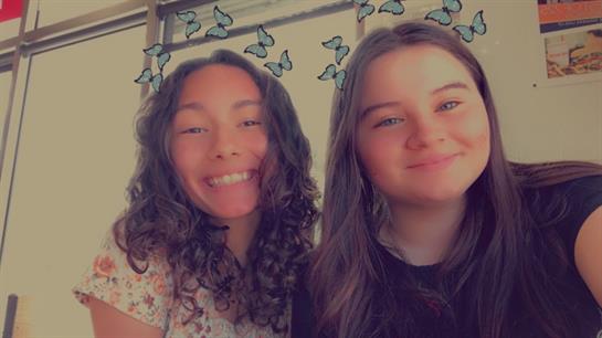 Two friends smile brightly while taking a selfie, surrounded by decorative butterflies, indoors.