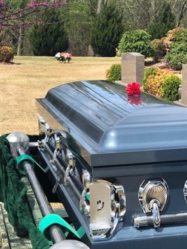 A casket is placed at a cemetery surrounded by lush green gardens under clear skies.