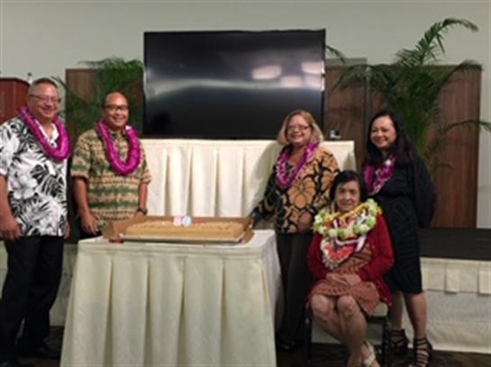 A group of smiling people wearing leis and traditional attire gathers around a table in celebration.
