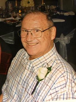 An elderly man smiles in a checked shirt and white rose at a festive gathering.