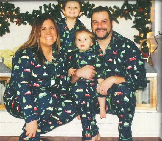 Family poses happily in matching pajamas with festive patterns by a decorated fireplace.