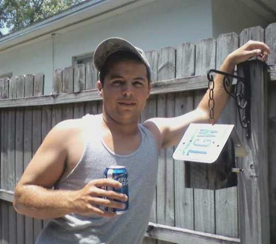 A man stands relaxed by a wooden fence holding a can, enjoying a warm day outdoors.