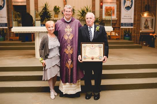 A couple stands proudly with a priest, holding a certificate in a church during their anniversary.