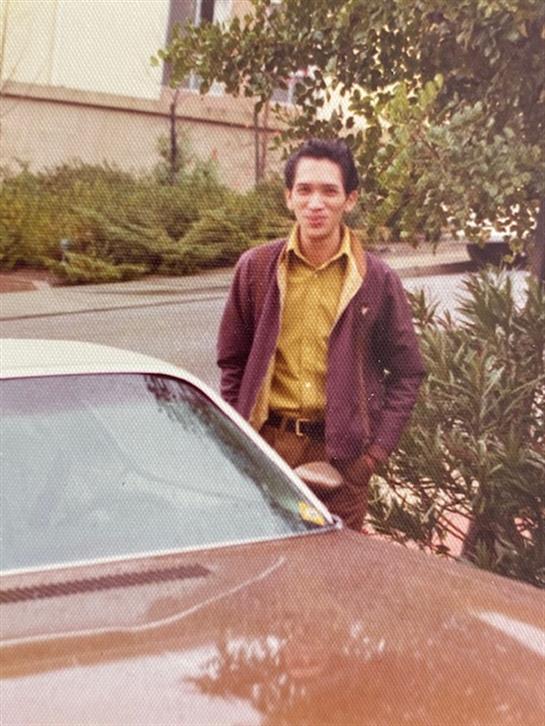 A young man stands beside a classic car on a city street, surrounded by greenery.