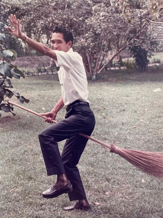 A man is enjoying a playful moment in a park while riding a broomstick, expressing joy.