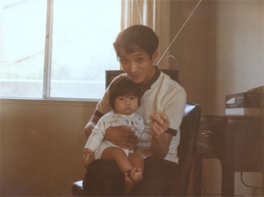 A young father sits on a chair, lovingly holding his baby in a softly lit room.
