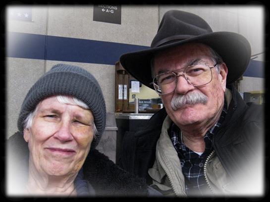 An elderly couple stands together, both smiling warmly while bundled up for cold weather.