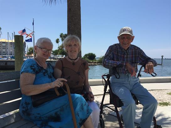 Three seniors sit together by the water, sharing stories and laughter under a clear blue sky.