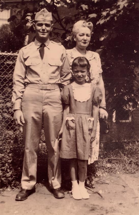 A soldier stands proudly next to a woman and a young girl in a garden, showcasing family ties.