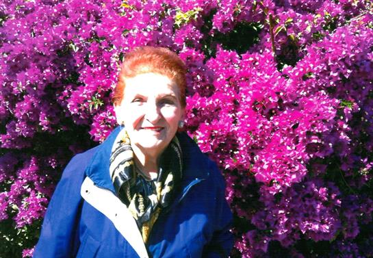 A cheerful elderly woman stands among blooming bougainvillea flowers, enjoying a bright day.