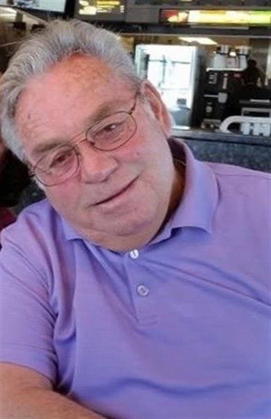 An elderly man with glasses smiles at the camera while sitting in a comfortable, casual atmosphere.